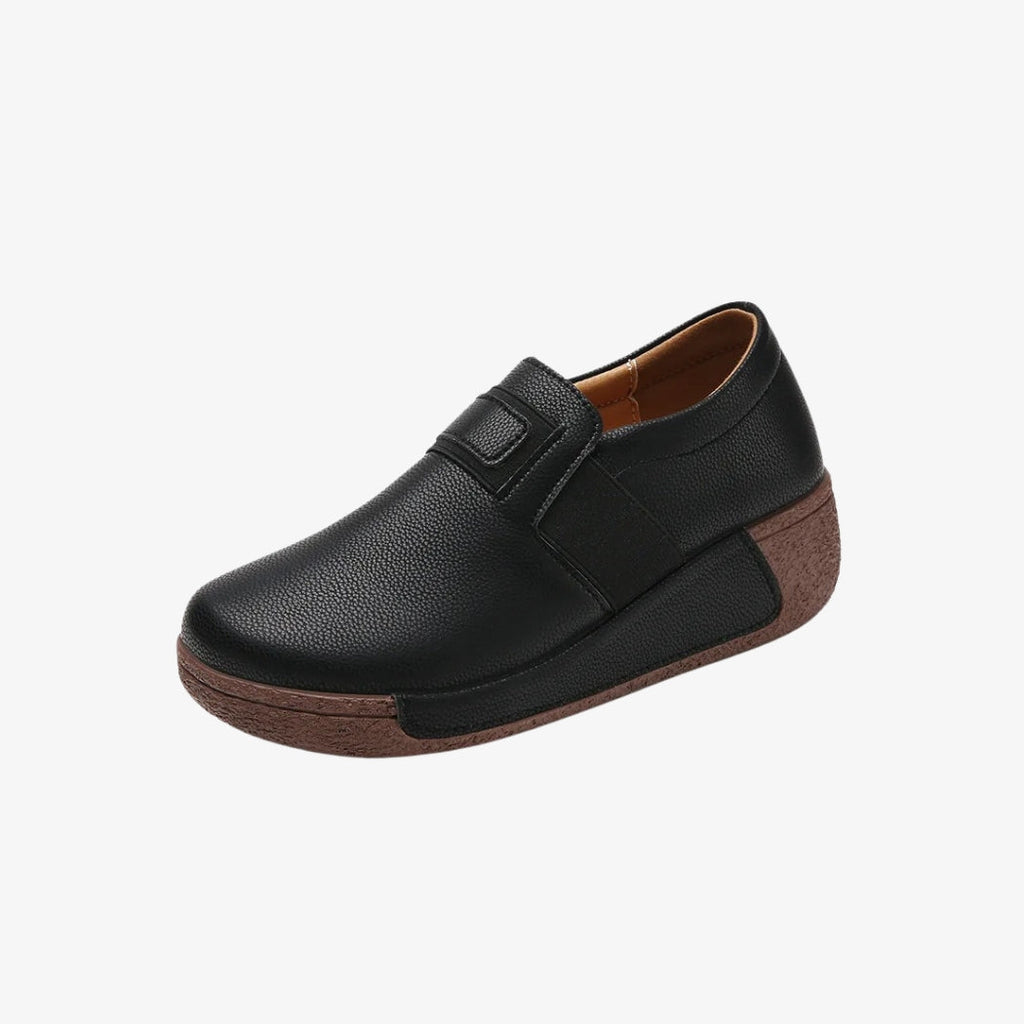 Ergonomiska kilklackloafers för dam i svart syntetiskt läder för optimal komfort. Skorna har en tjock, texturerad brun sula som ger ett utmärkt stöd. Den stilrena looken med elastisk öppning gör dem lätta att ta på samtidigt som de är eleganta och praktiska.