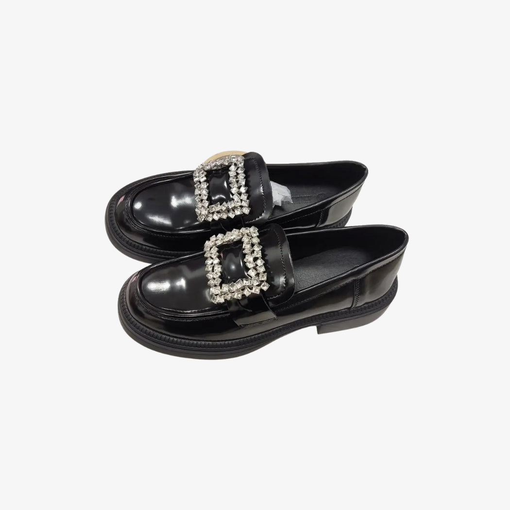 Svarta lackloafers för dam i syntetiskt läder, eleganta och blanka. Skorna har ett stort dekorativt spänne med gnistrande strass på ovansidan, som ger en glamourös och sofistikerad touch, perfekta för en chic outfit.