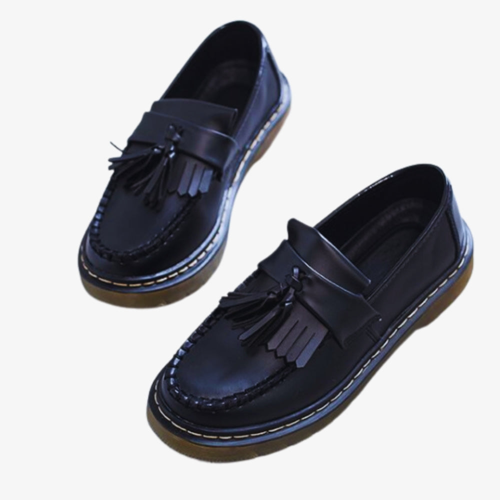 På en vit bakgrund syns ett par loafers med tofsar och fransar i syntetiskt läder för dam, de är svarta