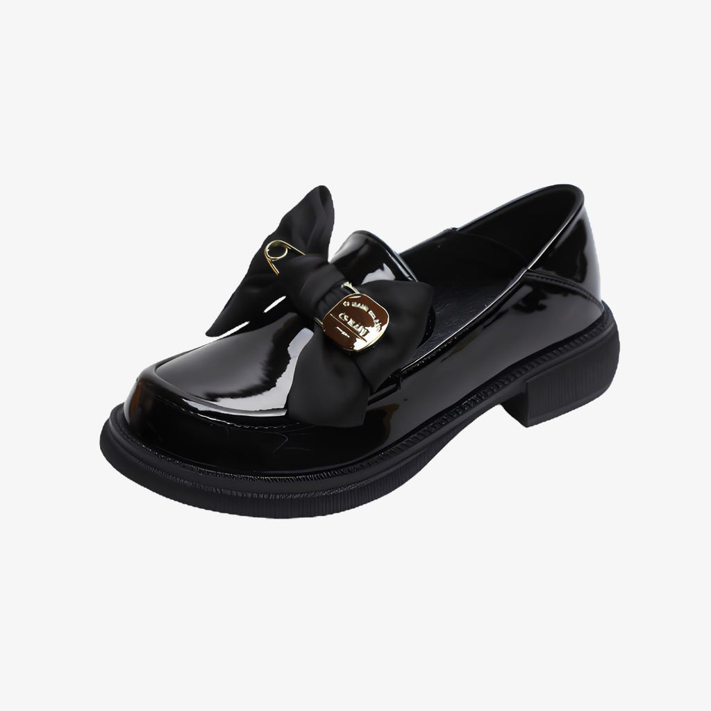 Svarta lackloafers med tjock sula, rosett och nål mot mycket ljusgrå bakgrund