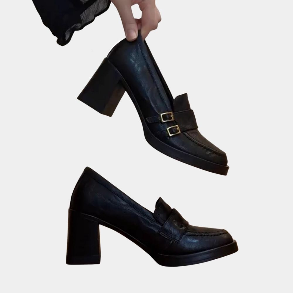 Ett par svarta högklackade loafers, där en sko står i profil och den andra hålls mellan fingrarna på en kvinna