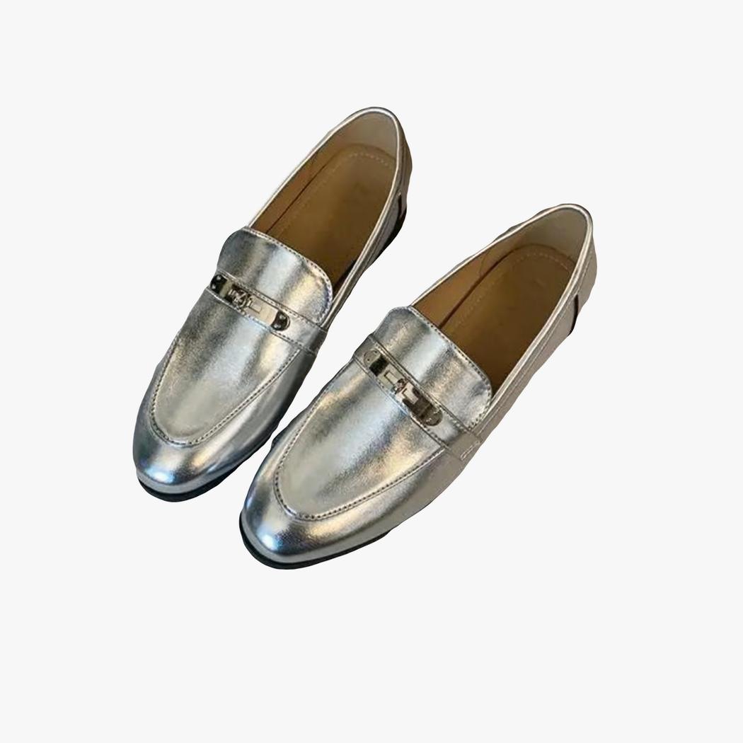På en ljus bakgrund syns ett par silverfärgade loafers med fyrkantig klack för dam