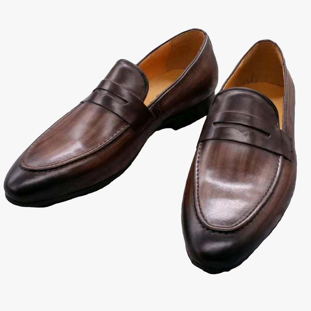 På en ljusgrå bakgrund syns ett par mörkbruna loafers för herr i äkta läder