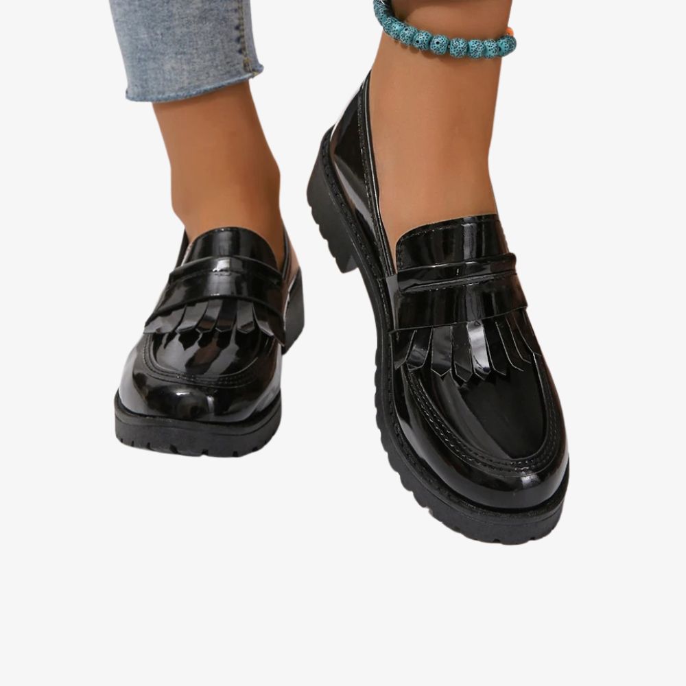Ett par svarta lackloafers med fransar burna av en kvinna i blå jeans och med fotlänk.