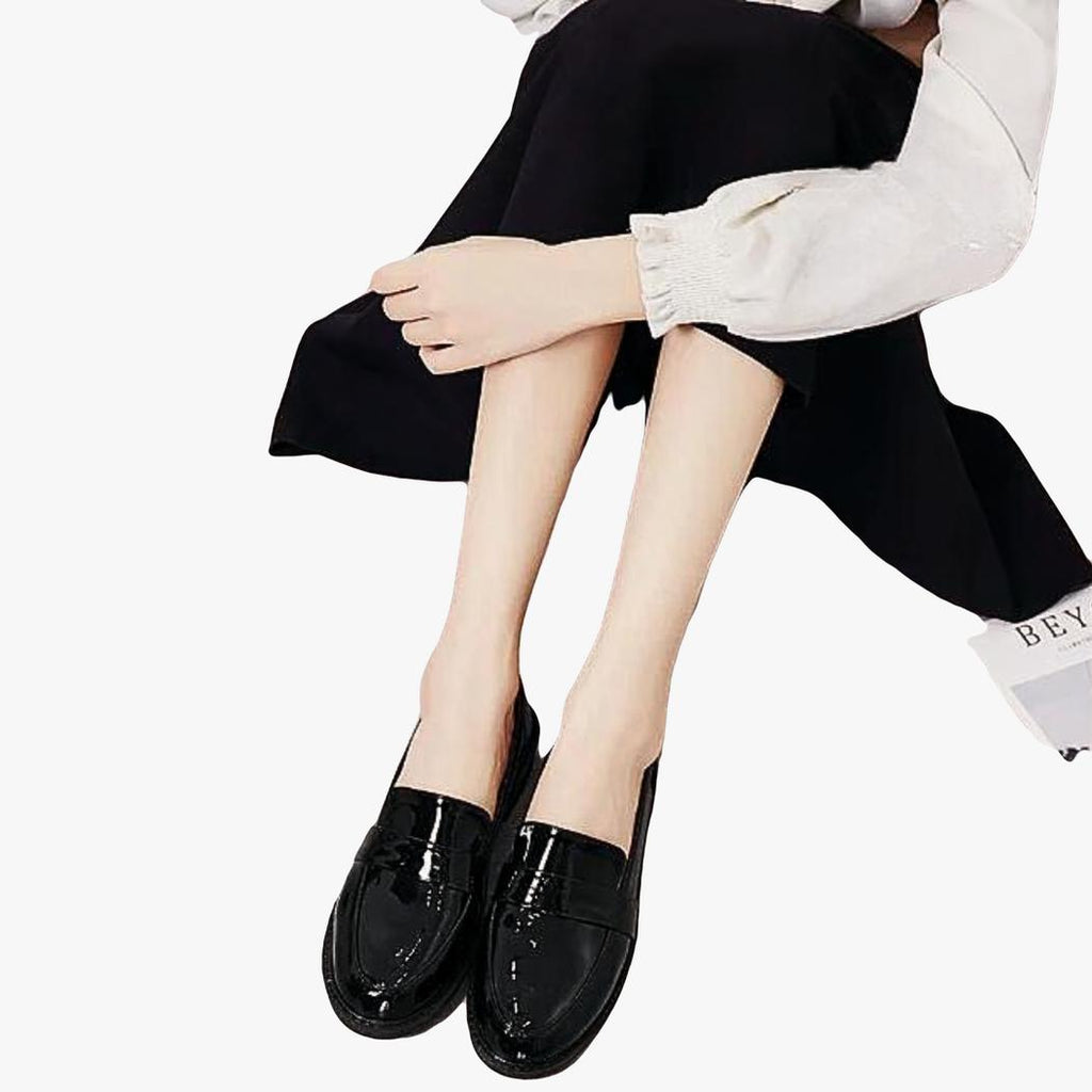 En kvinna sitter på golvet, överkroppen syns inte, och bär ett par svarta loafers med fyrkantig klack i preppystil