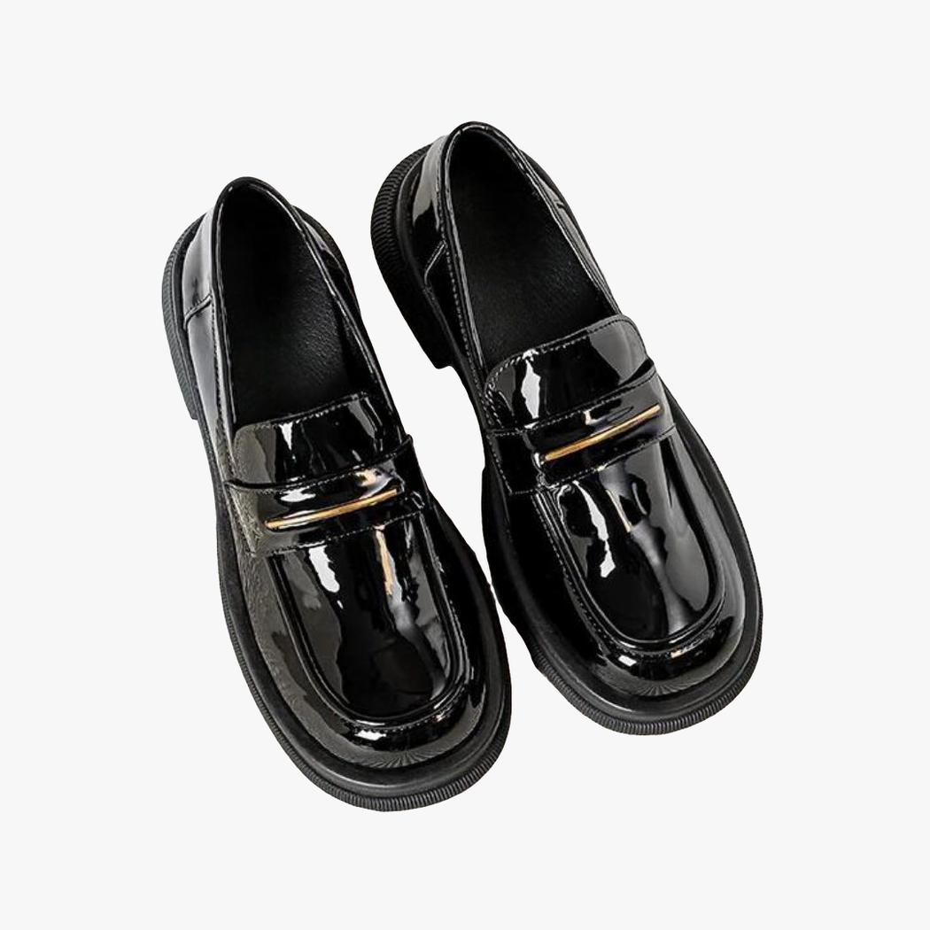 På en ljus bakgrund syns ett par svarta lackloafers i syntetiskt läder för dam