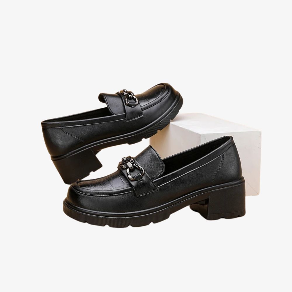 Ett par svarta loafers med klack, där en sko står på ett vitt block.
