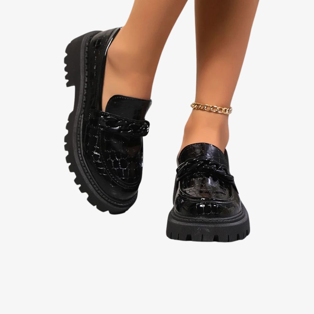 Ett par svarta lackloafers med krokodilmönster burna av en kvinna som syns från vristerna och nedåt. Hon bär en fotlänk.