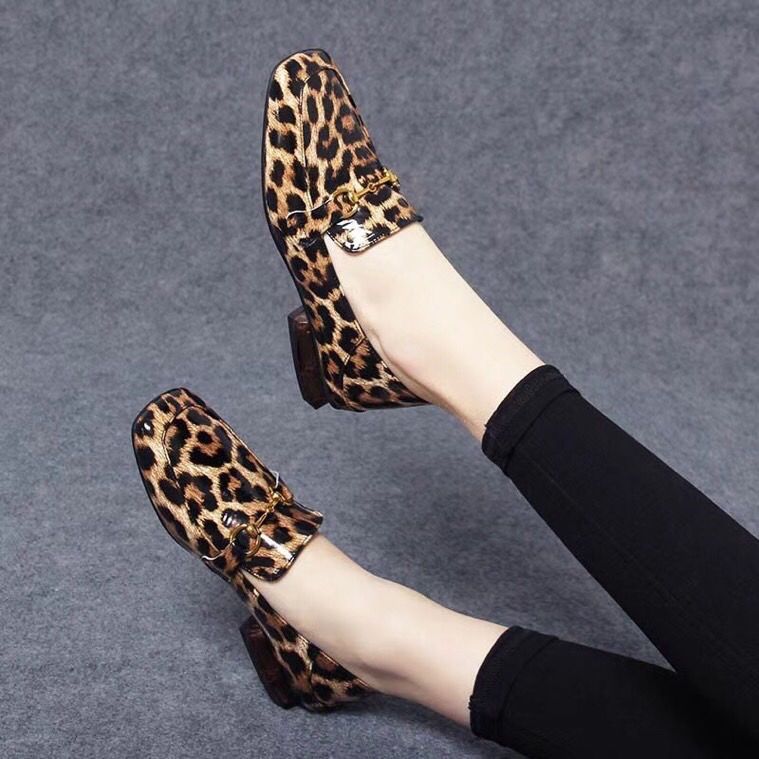 På en grå bakgrund, ett par ben med svarta leggings och blanka loafers med fyrkantiga klackar i leopardmönster