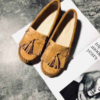 Indiska loafers i kamelfärgad mocka med fransar och tofsar, placerade på ett magasin på ett polerat betonggolv.
