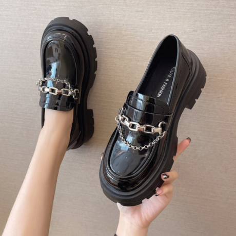 två svarta loafers i lackat konstläder med dekorativa kedjor framtill, tjock sula, den ena buren av ett ben, den andra buren av en hand, mot en beige väggbakgrund