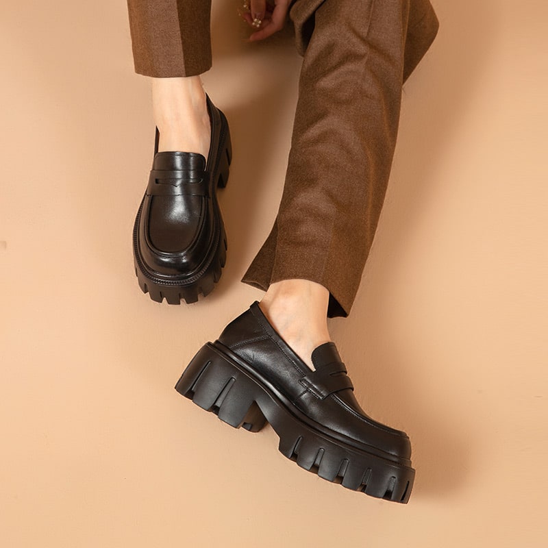 Foto av ett par svarta loafers som bärs av en kvinna med bruna byxor mot en rosa bakgrund
