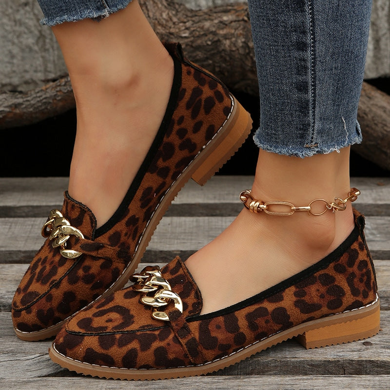Ett par bruna loafers med leopardmönster i profil, med en liten klack i träimitation och en guldkedja på ovansidan. På ena foten sitter en fotledskedja i guld.