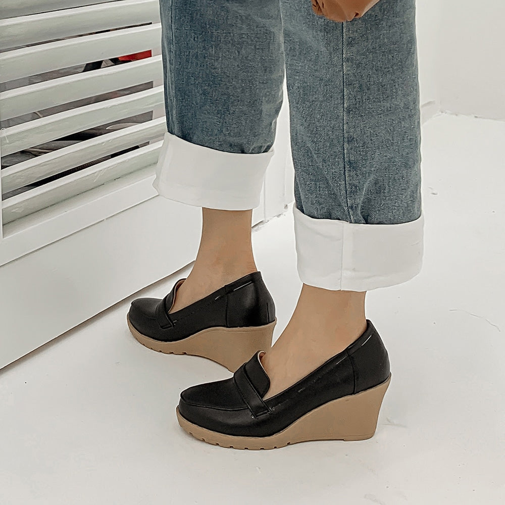 Foto av ett par svarta kilklackade loafers som bärs av en kvinna i jeans på ett grått golv