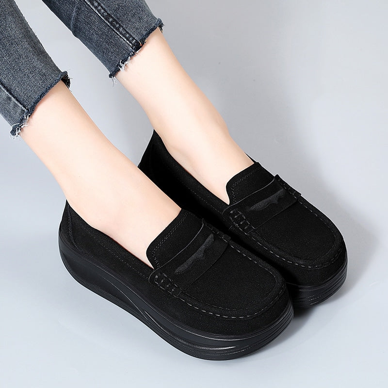 Foto av ett par svarta kilklackade loafers som bärs av en kvinna i jeans på ett grått golv