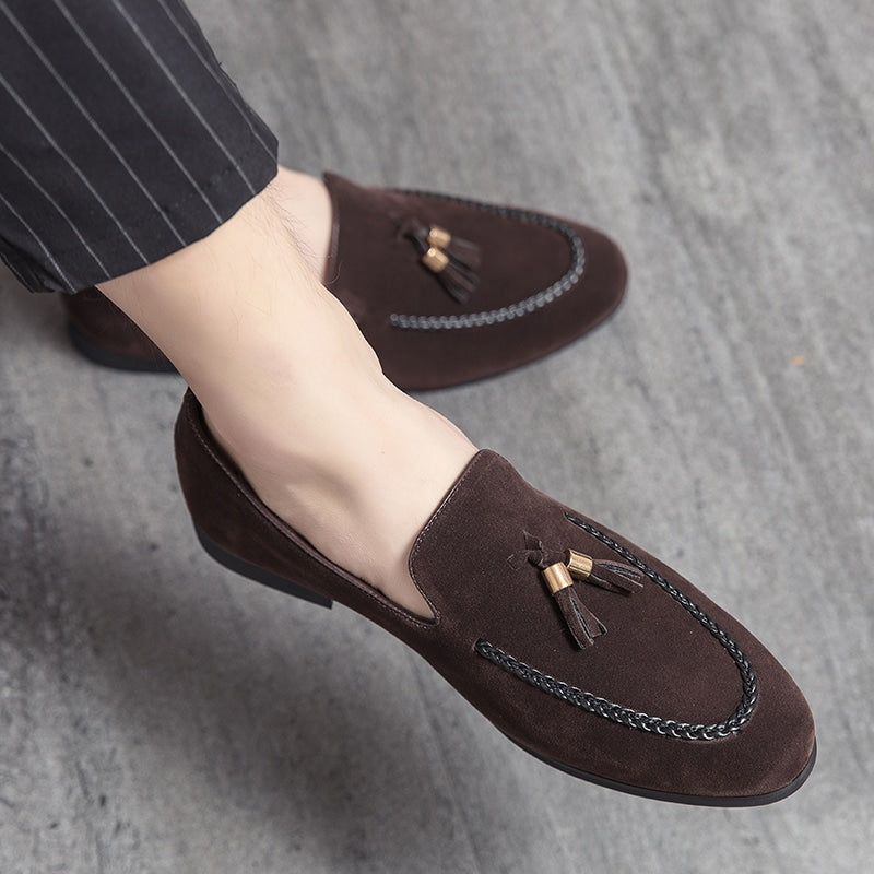 Mocka loafers med dekorativa tofsar och en läder twist på sömmen, vid fötterna på en korsbent man klädd i svarta kostymbyxor med tunna vita ränder.