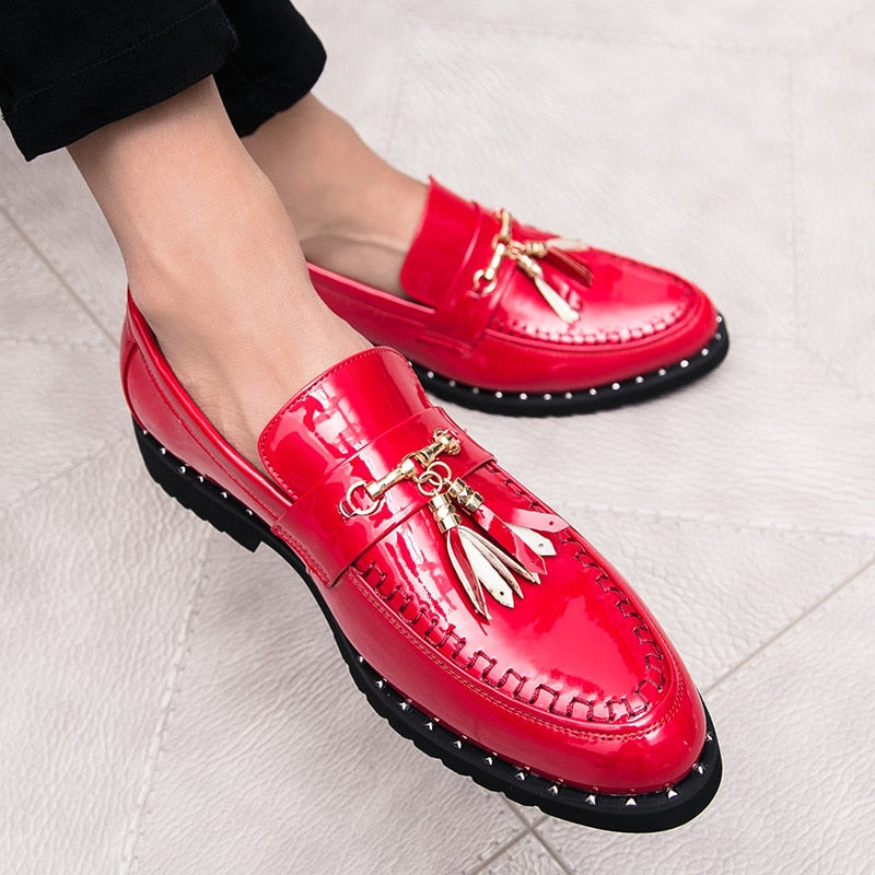 Ljusröda loafers i lack med dekorativa tofsar på fötterna av en man som sitter med benen i kors och bär svarta kostymbyxor.