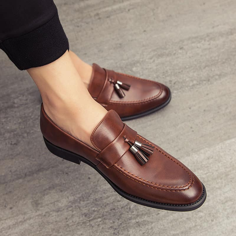 Bruna läderloafers med spetsiga tår och dekorativa tofsar på fötterna av en man som bär svarta byxor och korsade ben.