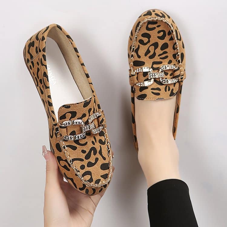 Ett par rundade loafers med brunt leopardmönster och silverfärgade strassringar, en sko på foten med svarta strumpbyxor och den andra i en hand