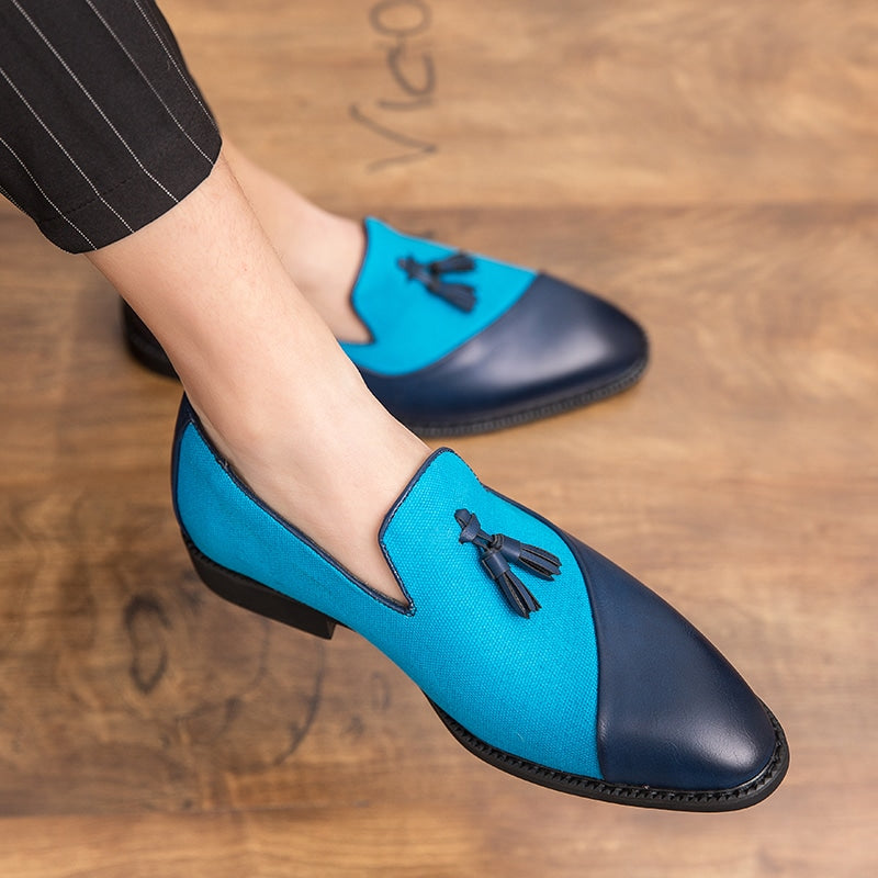 Mörk- och ljusblå loafers i läder och textil med dekorativa tofsar på fötterna på en man med korslagda ben som bär svarta kostymbyxor med fina vita ränder.