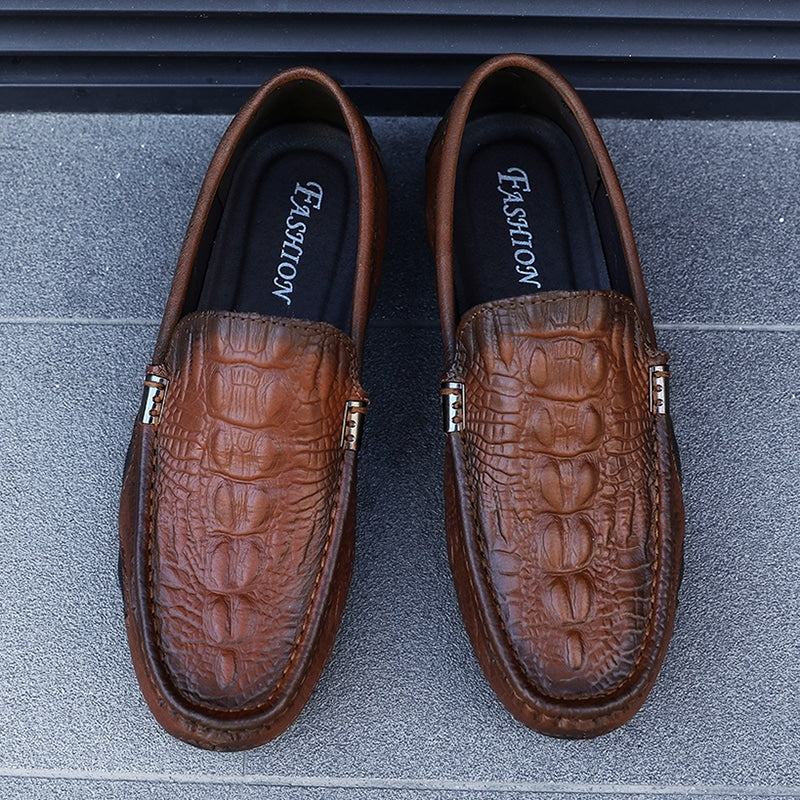 Bruna loafers med krokodilmotiv mot en grå bakgrund.