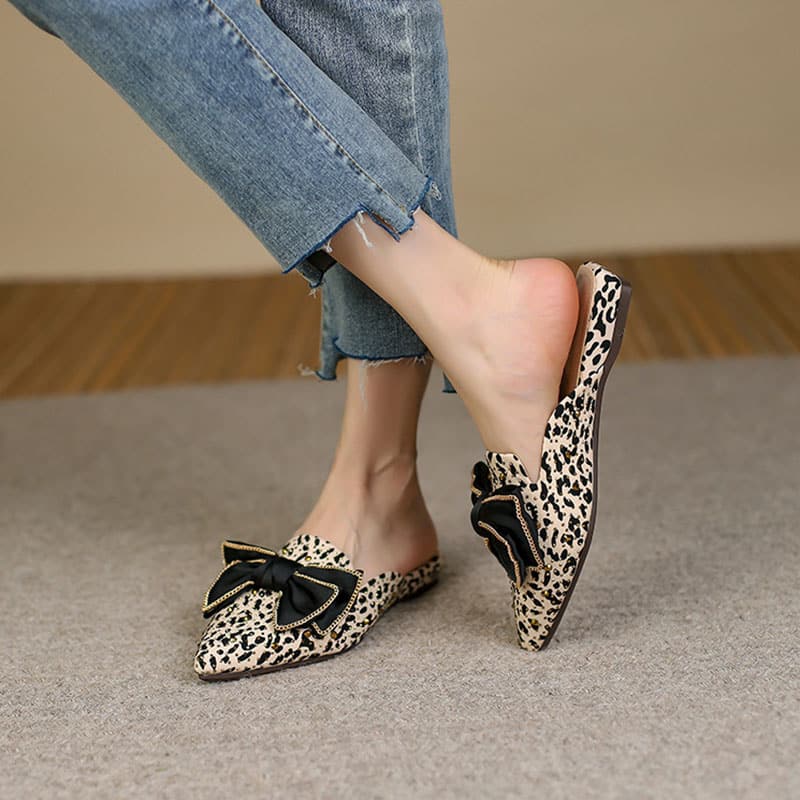 På en grå heltäckningsmatta ligger ett par spetsiga mockasiner med en svart rosett på toppen, leopardmönstrade och prydda med guldnitar. Paret bärs med korta, slitna blå jeans, anklarna är bara.