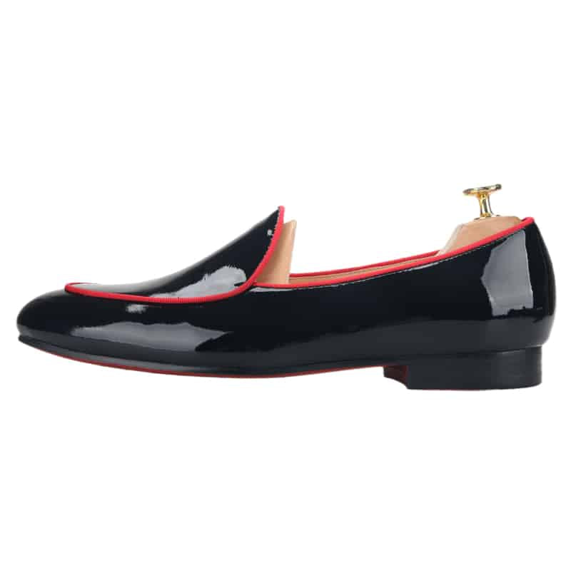 Svarta, blanka loafers med röd piping på tårna mot en vit bakgrund