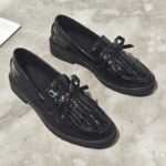 Ett par svarta loafers i lack med fransar på ett grått golv