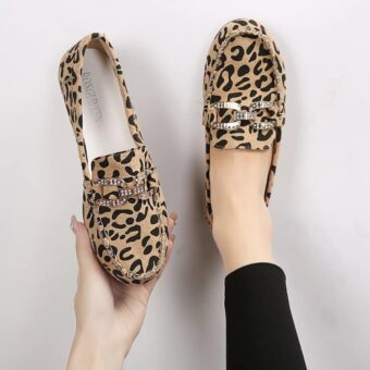 Ett par mockasiner i leopardmönster med sömmar och silverfärgade strassringar på ovansidan. En sko i ena foten och den andra i ena handen.