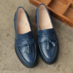 blå läderklackad loafer med tofs på toppen, placerad på grå mark