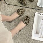 Ett par enkla loafers med leopardmönster från ovan bärs med en vit kjol och bara fötter. Golvet är en beige matta och det ligger en vit tidning bredvid