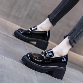 Svarta lackade loafers med klack och tjocka svarta sulor på fötterna av en kvinna som står i svarta skinny jeans.