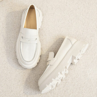 Ett par enkla vita loafers i slätt läder med tjocka vita sulor på ett ljusbeige golv.