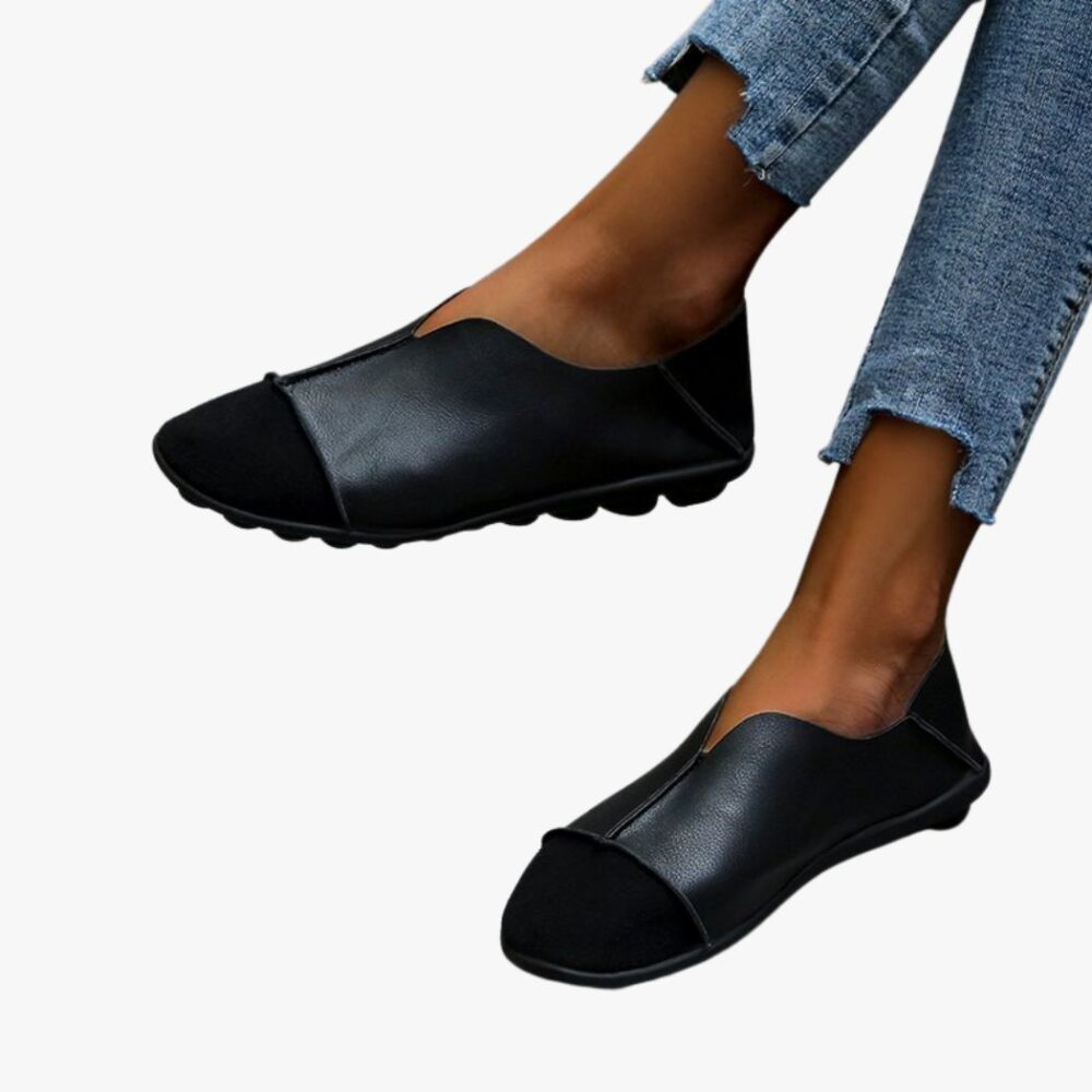 Ett par mjuka loafers med rund tå i mocka och slätt läder med synliga sömmar som bärs med jeans på ett ljusgrått klinkergolv