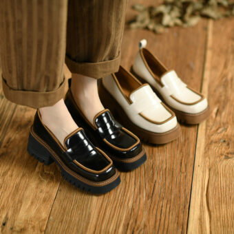 Två par loafers i kontrasterande lackskinn, den ena beige och brun, den andra svart och brun, vid fötterna på en kvinna som står på ett parkettgolv och bär breda bruna randiga byxor.